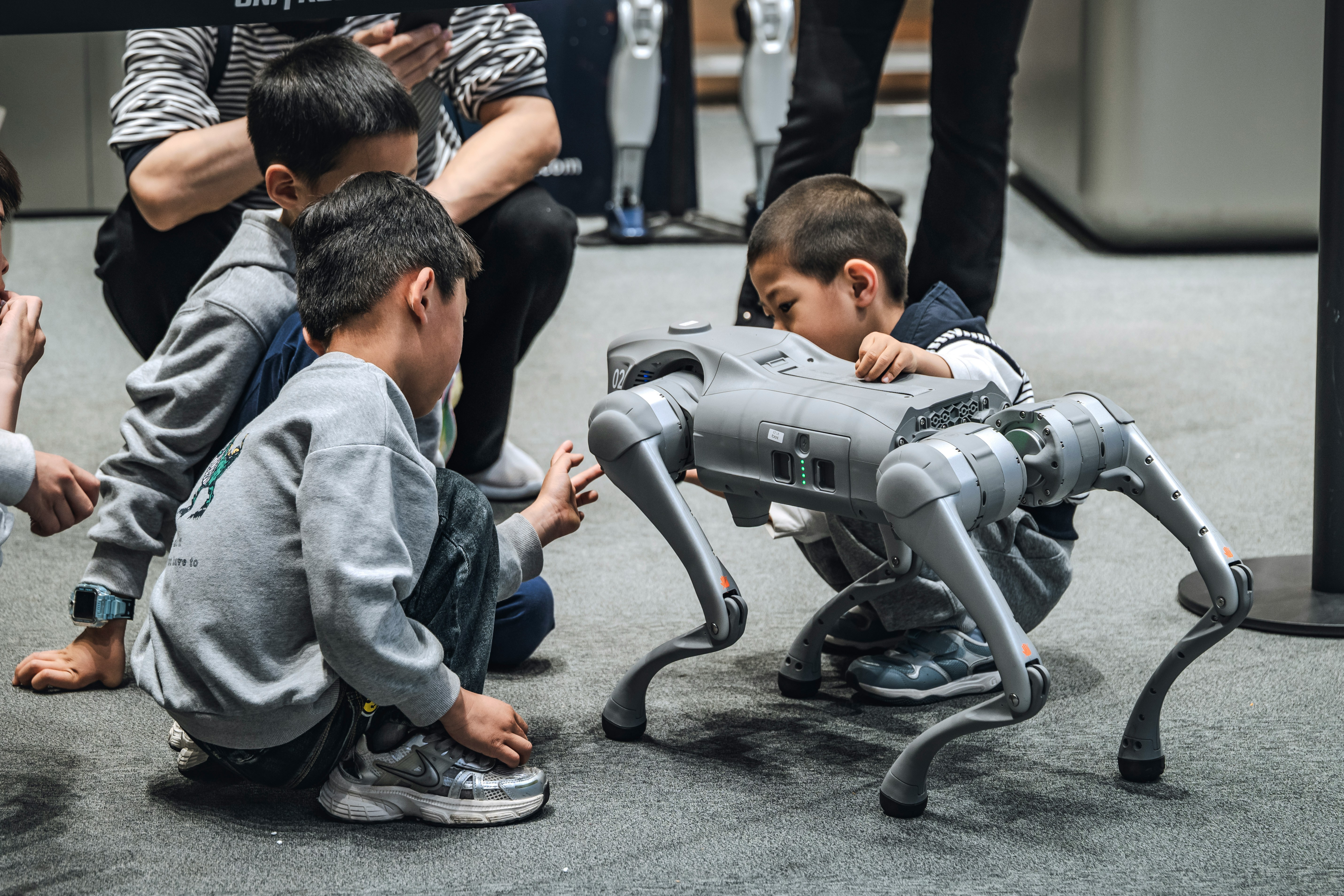 Children crouch around a large quadruped robot dog, touching its legs and observing it with curiosity in an indoor exhibition space