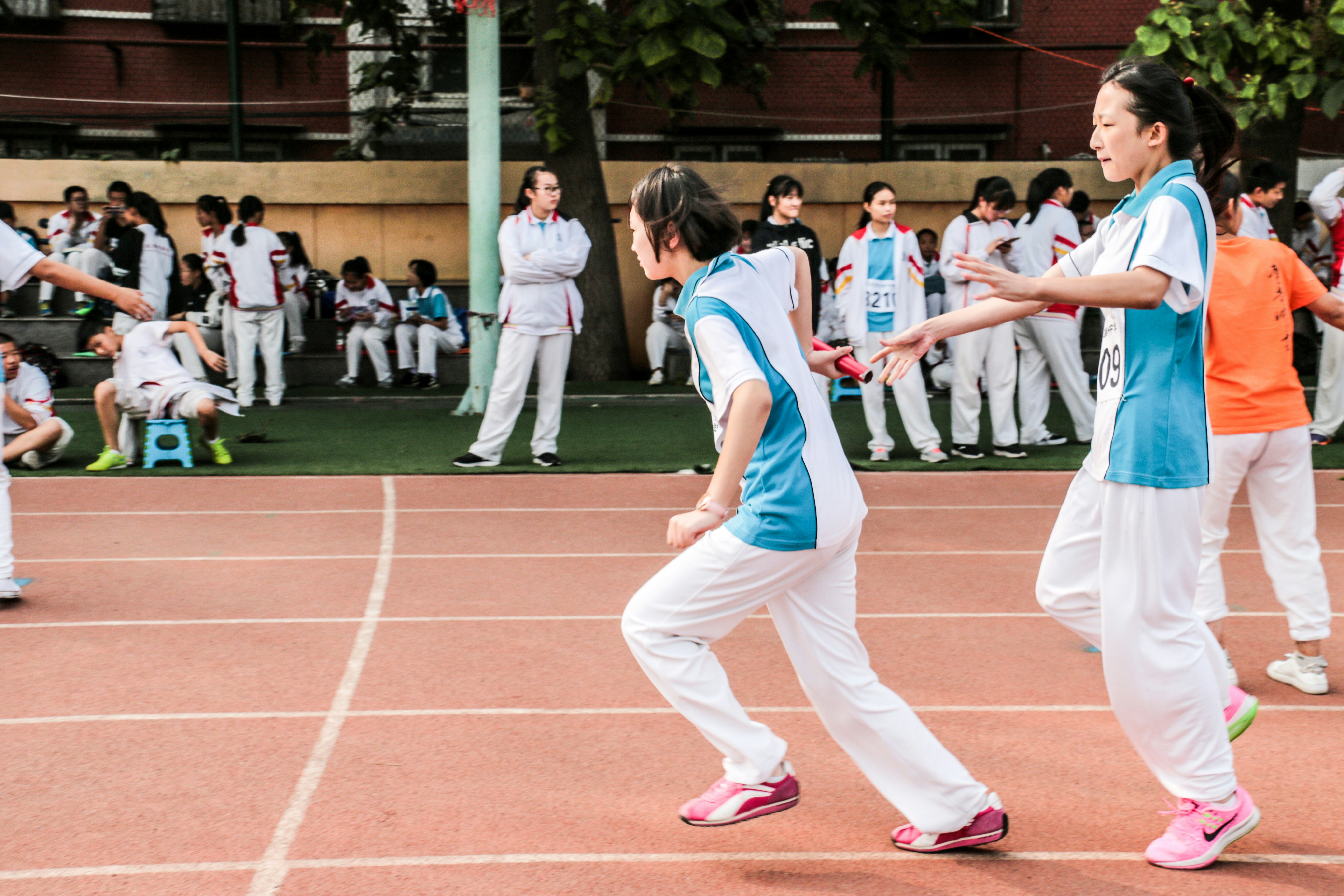 Students in blue-and-white sports uniforms move across a red track during a lively school athletics event, with classmates watching.