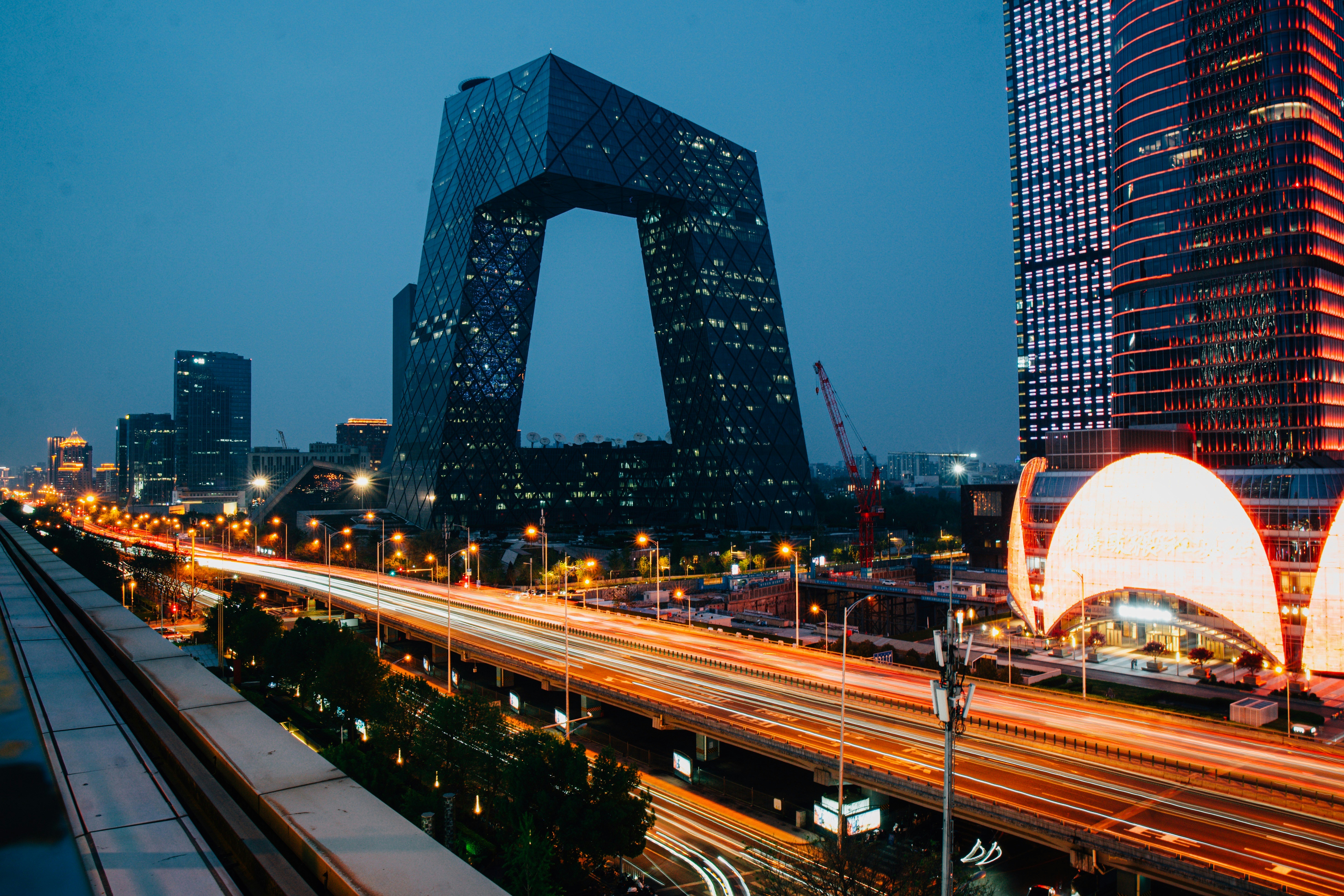 Nighttime cityscape with Beijing’s CCTV headquarters, glowing highways, modern towers, and a bright dome-shaped building under a blue sky.